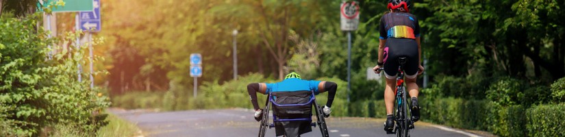 Bild zeigt einen Rollstuhlfahrer neben einem Fahrradfahrer von hinten auf leerer Straße, die durch ein Wald führt.