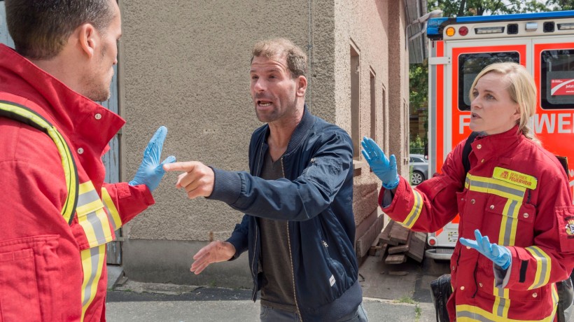 Das Bild zeigt eine angespannte Situation in einem Rettungswagen. Zwei Rettungskräfte in roten Uniformen mit reflektierenden Streifen stehen einem Mann gegenüber, der scheinbar aggressiv oder unkooperativ wirkt. Der Mann wird von einem der Rettungskräfte an den Schultern gehalten, vermutlich um ihn zu beruhigen oder zurückzuhalten. Die andere Rettungskraft, eine Frau, schaut aufmerksam und besorgt auf die Situation. Das Bild zeigt eine angespannte Situation in einem Rettungswagen. Zwei Rettungskräfte in roten Uniformen mit reflektierenden Streifen stehen einem Mann gegenüber, der scheinbar aggressiv oder unkooperativ wirkt. Der Mann wird von einem der Rettungskräfte an den Schultern gehalten, vermutlich um ihn zu beruhigen oder zurückzuhalten. Die andere Rettungskraft, eine Frau, schaut aufmerksam und besorgt auf die Situation.