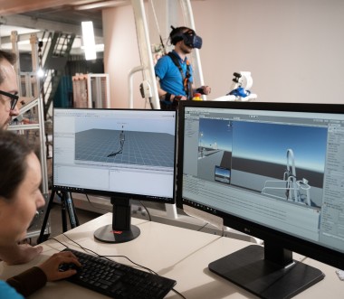 Zu sehen sind zwei Personen vor zwei Bildschirmen, die sich eine Gang-Computersimulation anschauen. Im Hintergrund sieht man die eine Testperson in einem Ganzkörpermessanzug auf einem Laufband. Zu sehen sind zwei Personen vor zwei Bildschirmen, die sich eine Gang-Computersimulation anschauen. Im Hintergrund sieht man die eine Testperson in einem Ganzkörpermessanzug auf einem Laufband.