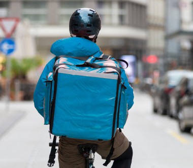 Die Person trägt einen dunklen Helm, sowie eine Jacke und einen Lieferdienst-Rucksack im gleichen hellblauen Farbton. Die Person trägt einen dunklen Helm, sowie eine Jacke und einen Lieferdienst-Rucksack im gleichen hellblauen Farbton.
